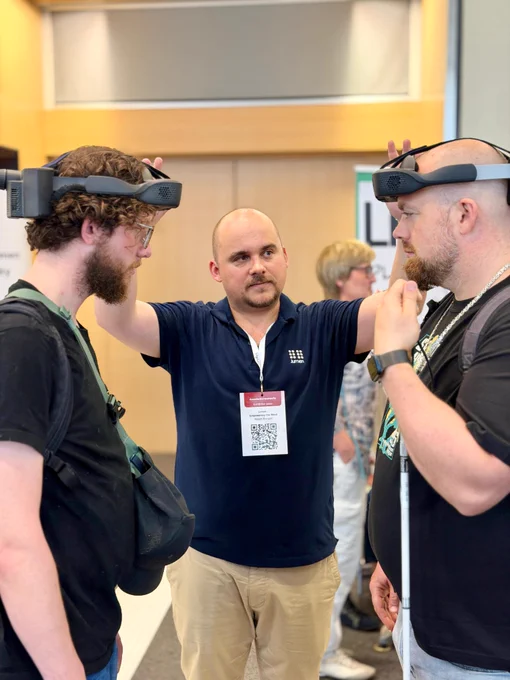 Balázs Blényesi, .lumen’s Chief Business Development Officer, stands between two visitors at SightCity in Frankfurt, guiding a double demo of the .lumen Glasses. Both visitors are wearing the headsets as Balázs explains the experience, creating a focused, hands-on moment on the exhibition floor.