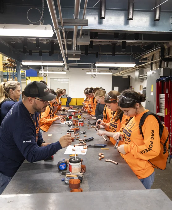 Alt text: Students in bright orange shirts work with copper piping and tools during a hands-on trades workshop led by an instructor.