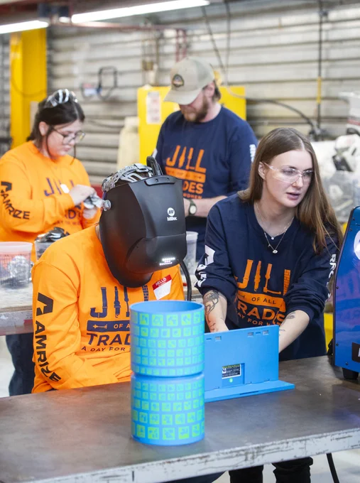 Alt text: Students wearing safety glasses and bright orange “Women of All Trades” shirts participate in a hands-on welding and trades activity in a workshop classroom. One student wears a welding helmet while another assists beside a blue welding machine.