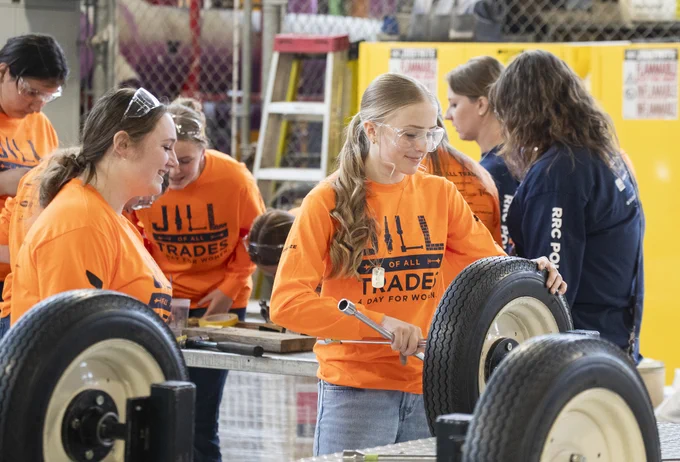 Alt text: Students wearing bright orange “Women of All Trades” shirts and safety glasses work together on a hands-on tire and mechanics activity in a workshop.