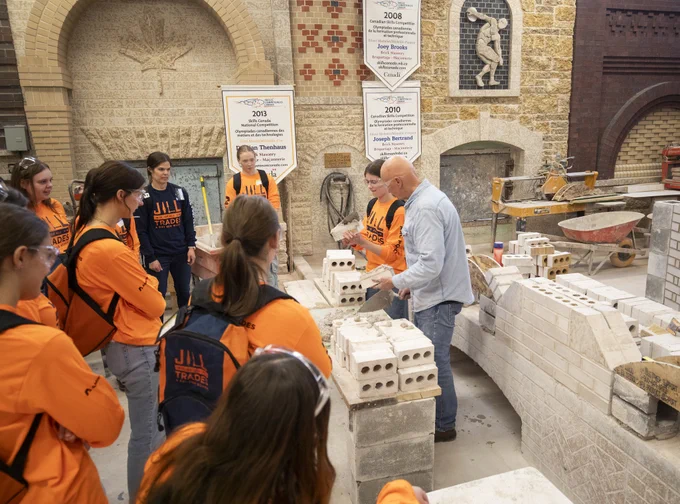 Alt text: Students wearing bright orange “Women of All Trades” shirts gather around an instructor demonstrating bricklaying techniques in a masonry workshop filled with bricks, tools and partially built walls.
