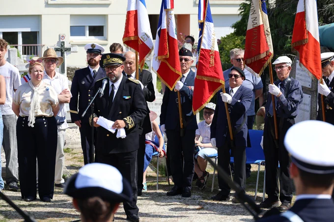 Un groupe de personnes, dont plusieurs en uniforme militaire et officiel, se tient debout dans un cimetière lors d’une cérémonie commémorative. Certaines personnes tiennent des drapeaux, et l’atmosphère est solennelle. En arrière-plan, on aperçoit plusieurs tombes et croix. La scène a lieu en plein jour, sous un ciel ensoleillé.