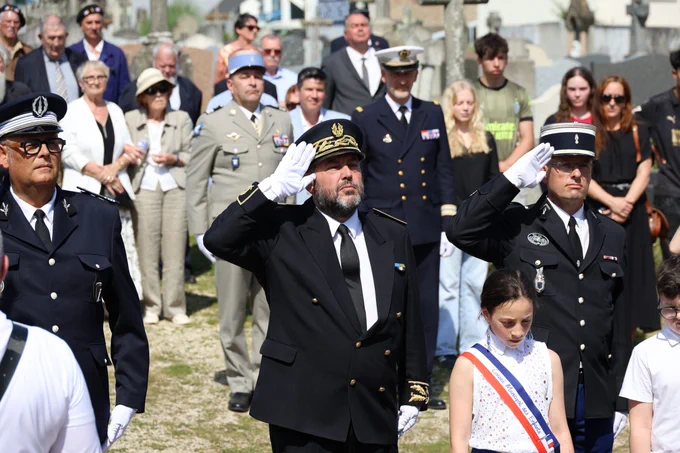 Cérémonie commémorative en extérieur, avec plusieurs officiers en uniforme militaire et policier au premier plan, saluant solennellement. Au centre, un homme en uniforme noir avec une casquette dorée. À côté de lui se tient une jeune fille portant une écharpe tricolore. En arrière-plan, un groupe de civils, hommes et femmes, observe la scène. L'événement se déroule sous le soleil, dans un cimetière à Concarneau.
