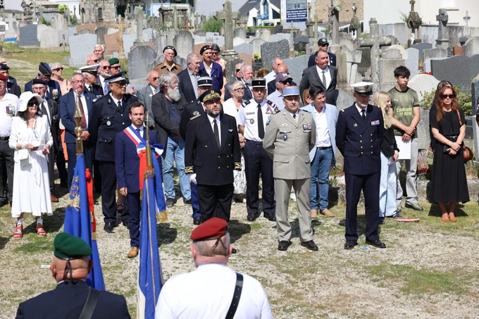 Un groupe de personnes, dont plusieurs en uniforme militaire et officiel, se tient debout dans un cimetière lors d’une cérémonie commémorative. Certaines personnes tiennent des drapeaux, et l’atmosphère est solennelle. En arrière-plan, on aperçoit plusieurs tombes et croix. La scène a lieu en plein jour, sous un ciel ensoleillé.
