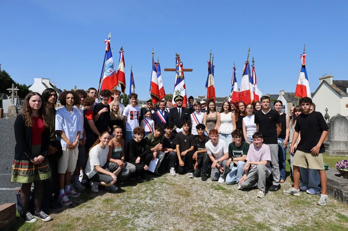 Un grand groupe de jeunes et de quelques adultes posent ensemble dans un cimetière sous un ciel bleu. Derrière eux, plusieurs personnes en uniforme tiennent des drapeaux tricolores français décorés de rubans, et une croix en bois est visible à l’arrière-plan. Les bâtiments et des tombes entourent la scène, évoquant une cérémonie de commémoration.