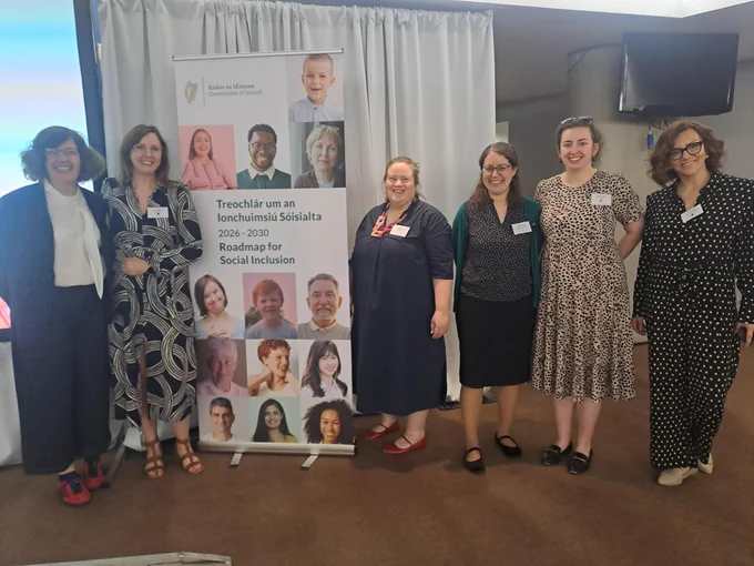 A group of seven people pose together at an indoor event, smiling at the camera. They stand beside a Government of Ireland roll-up display banner promoting the Roadmap for Social Inclusion 2026–2030, which features a grid of portrait photographs of diverse individuals. Several attendees are wearing name badges. A projection screen is visible to the left and a TV monitor to the upper right.