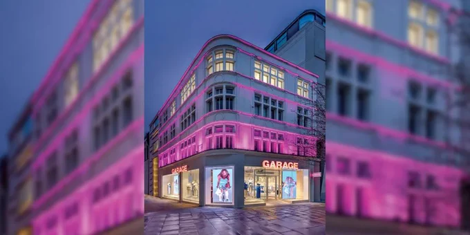 Corner view of a multi-story store with pink exterior lighting and "GARAGE" signs, displaying clothing in large glass windows at dusk.