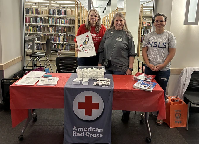 Three volunteers stand behind a Red Cross table in front of a library entrance