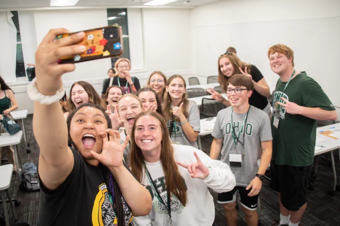 A group of people smiling and gesturing while posing for a selfie in a classroom. Several are wearing shirts with "Ohio University" logos.
