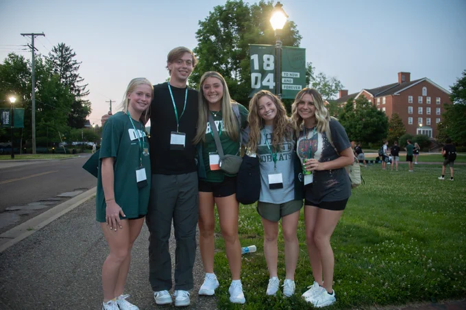 A group of five people smiling outdoors, standing near a light pole with "1804" and "Ohio" banners. They are wearing casual attire and lanyards, suggesting a social or college event. A red brick building is in the background.