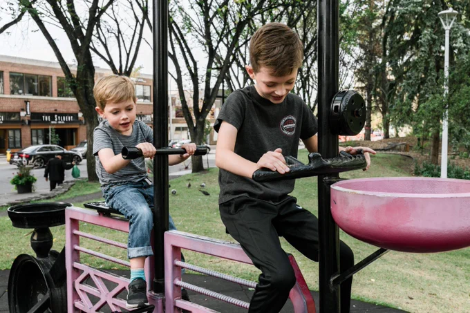 Two boys playing on a pink and black outdoor exercise machine in a park with trees and buildings in the background.