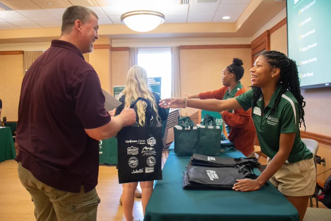 A person at a registration table hands a tote bag to a visitor. Other tables and attendees are visible in the background, creating a welcoming event atmosphere.
