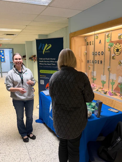 Two women stand near a mental health awareness table with informational materials in a school hallway.