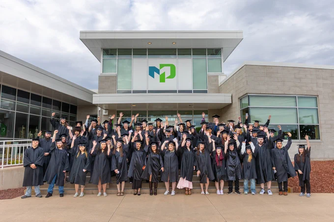 A large group of graduates in black caps and gowns stand outside a modern school building for a class photo. Many of the graduates have their hands raised in celebration beneath a large school logo on the building.