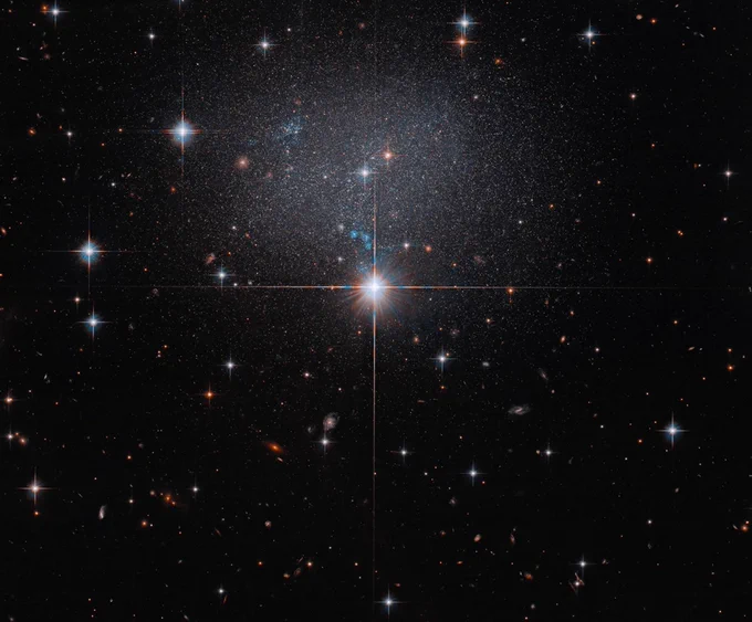 A foreground star at image center shines brightly with four diffraction spikes. Above this star is a diffuse and faint dwarf irregular galaxy. The galaxy itself has bright blue and red stars.