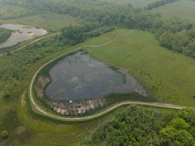 Aerial view of a heart-shaped pond surrounded by greenery and a winding dirt path in a lush, wooded area.