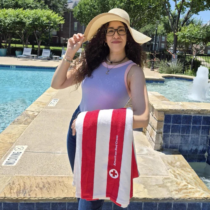 A person standing by a pool holds a red and white towel with the American Red Cross logo. They are wearing a wide-brimmed hat and glasses, smiling at the camera. There are trees and poolside chairs in the background.