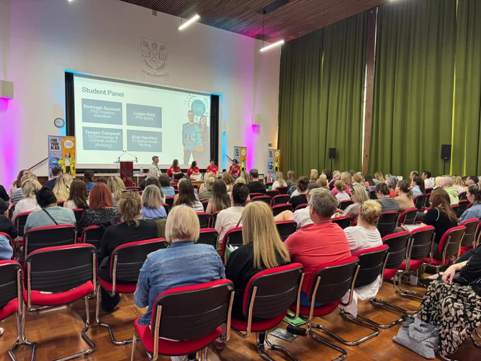 A large group is seated in a room facing a student panel presentation. The presentation slide includes names: Keegan Stewart, Logan Kiwi, Tegan Campbell, and Ben Riddington. The room features a display screen and green curtains.