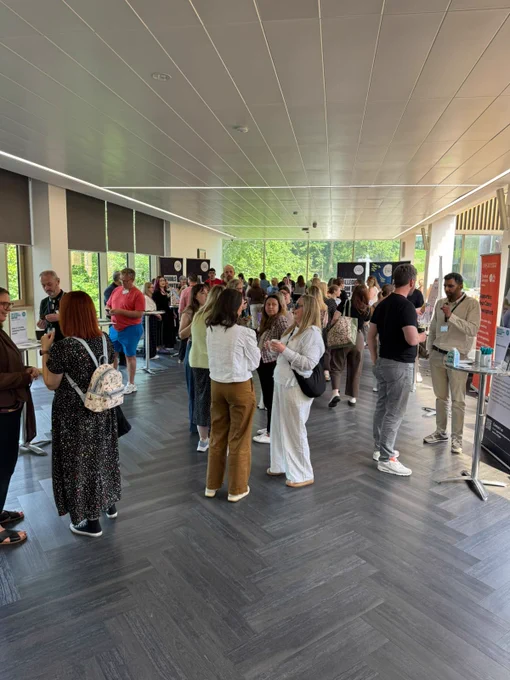 A group of people are gathered in a bright, modern hallway. Some are engaged in conversation while others stand or walk around. The space features large windows, allowing natural light to illuminate the room. There are promotional banners and informational stands visible. The atmosphere appears to be social and professional.