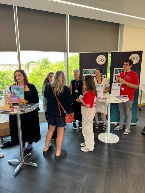 A group of people are engaged in conversation at an indoor event. Two booths display banners with "UWS" logos. One person is holding a booklet with colorful charts. The room has large windows with views of green trees outside.