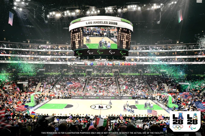 A vibrant basketball game during the Los Angeles 2028 Paralympics is taking place in a large indoor arena filled with spectators. The scoreboard displays the event details, and the court features players in wheelchairs. The arena is decorated with flags, and there is a noticeable presence of green lighting effects. Logos for LA28 and the Paralympics are visible at the bottom. This is an artistic rendering of what the venue can look like