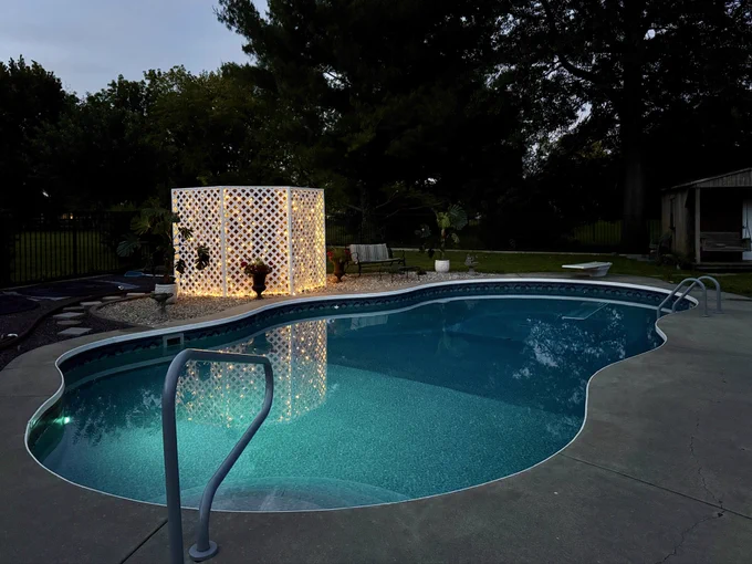 Our backyard in-ground swimming pool at dusk with clear turquoise water, featuring a newly built by me white lattice pool enclosure illuminated with warm string lights. The lights create a beautiful reflection in the pool, with a concrete deck, handrails, and landscaped backyard visible in the background.