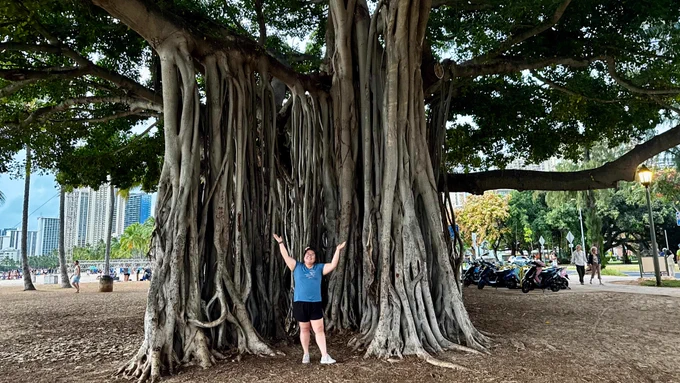 Brenda Such standing with her arms raised, surrounded by Hawaii’s Banyan tree.