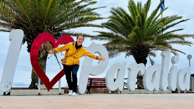 Katrina Pineda in Greece, posing and laughing in front of a word sculpture that reads, I heart Paralia.