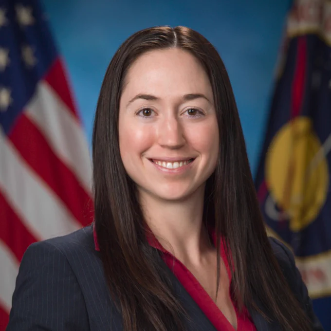 CALS alumna Grace Douglas wearing a navy blazer and red blouse, standing in front of American and NASA flags.