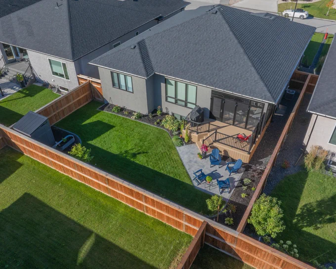 Aerial view of a landscaped backyard featuring a deck, stone patio with seating, screened-in sunroom, green lawn, and surrounding wooden fence.