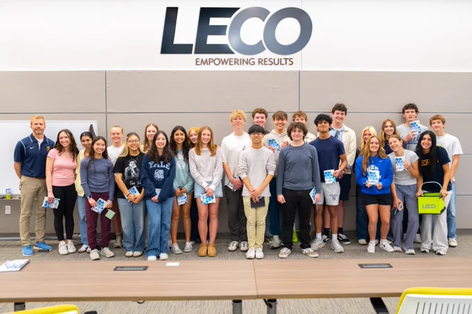 Group of diverse students and a teacher standing in front of a LECO sign in a classroom setting.