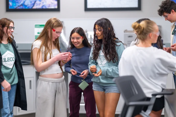 A group of young people standing and chatting casually in a modern indoor setting with screens on the wall.