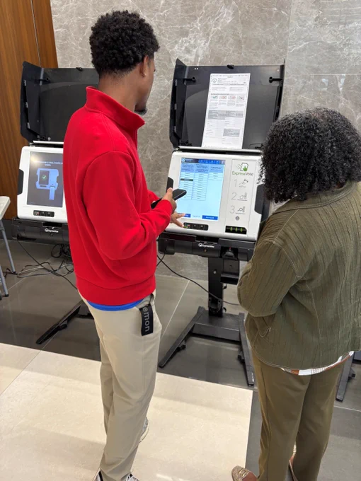 Two people are standing at electronic voting machines, learning about ranked choice voting. One person in a red jacket is interacting with the screen, while the other observes. 