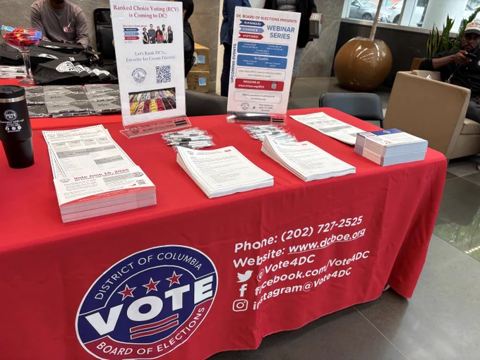 This image shows a table covered with a red cloth featuring the "District of Columbia Board of Elections" logo. The table holds informational materials, including flyers and brochures about ranked choice voting and a webinar series. Contact information for the D.C. Board of Elections is displayed on the tablecloth, along with various social media handles. 