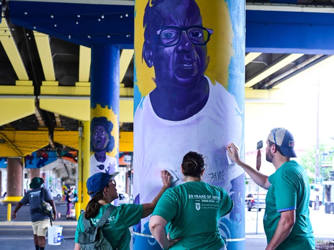 A group of Tulanians wearing green shirts are painting a mural on a large pillar under a bridge. The pillar features a portrait of a person wearing glasses. More art can be seen on surrounding pillars. The setting is urban with a bright and colorful atmosphere.