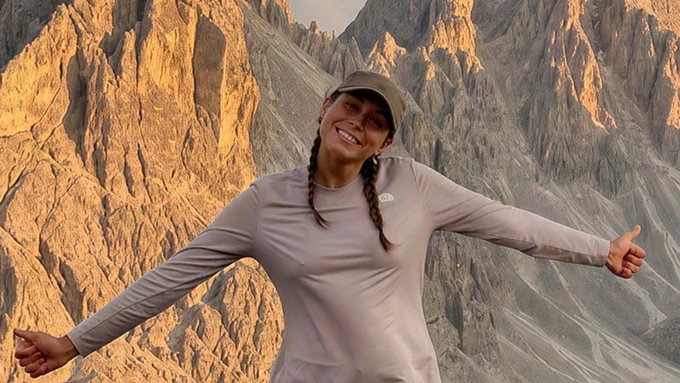 Science graduate Dria McKenna is smiling with arms outstretched in front of a rugged, sunlit mountain landscape. She is wearing a cap and a long-sleeved shirt.