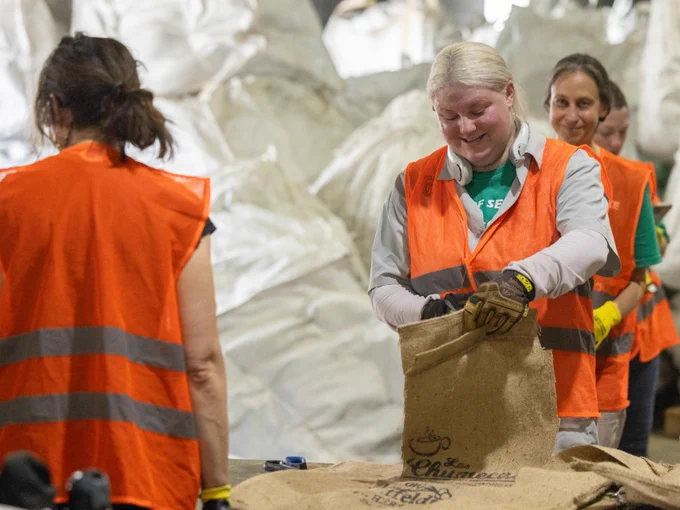 A group of Tulane staff wearing orange safety vests are working together, handling burlap sacks in what appears to be an industrial or warehouse setting. One person in the foreground is focused on securing the top of a sack. Large white bags are stacked in the background.