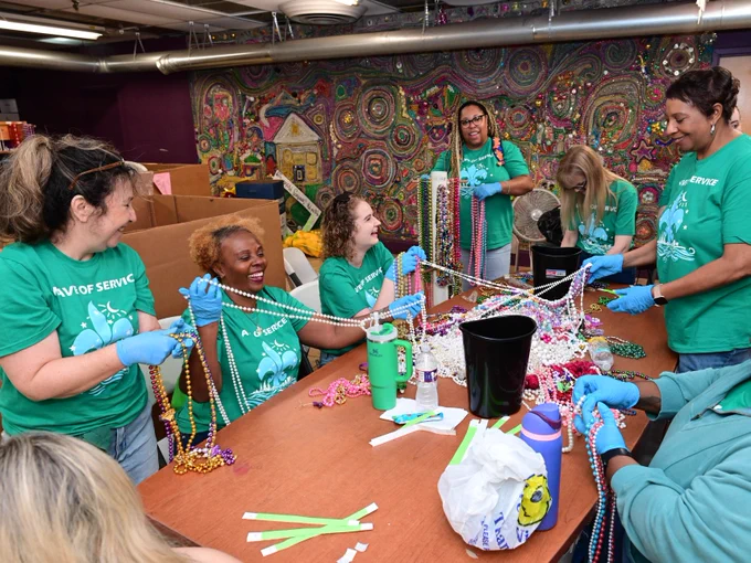 A group of people wearing matching green "Day of Service" shirts are gathered around a table, sorting colorful Mardi Gras beads. They are smiling and working together in a room decorated with vibrant, textured wall art depicting a house and swirling patterns.