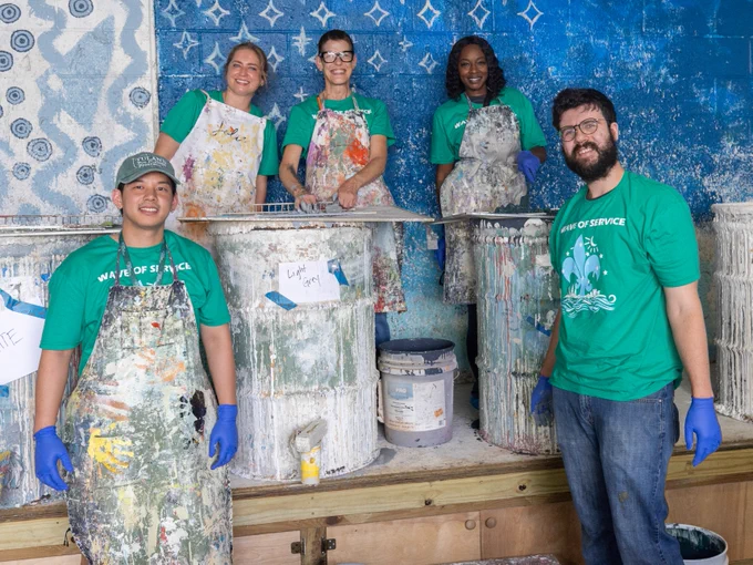 A group of five people, wearing matching green "Wave of Service" shirts and paint-splattered aprons, are smiling. They are standing in front of large paint containers, seemingly engaged in a painting or art project. The background features a blue and white patterned wall.