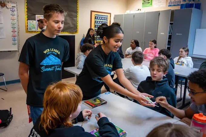 Students wearing WEB Program T-shirts help younger students in a classroom setting.