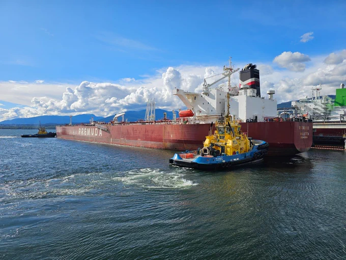 Large red cargo ship docked with a yellow tugboat assisting in calm harbor waters under a blue sky.