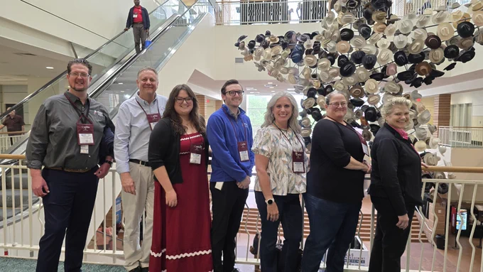 A group of seven people stand in a line indoors, smiling at the camera. They are in front of a modern art installation composed of numerous hanging hats of various colors. An escalator with a person descending is visible in the background. The setting seems to be a spacious, well-lit building.