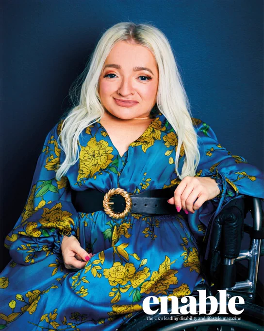 A professional photo portrait of Samantha Renke. She is smiling, has long blond hair and is wearing a blue silk floral dress and a black belt with gold clasp. She is using a wheelchair.