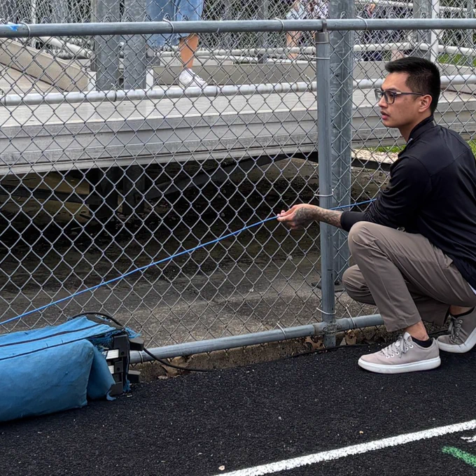 Tech worker crouching by a chain-link fence holding a blue video cable.