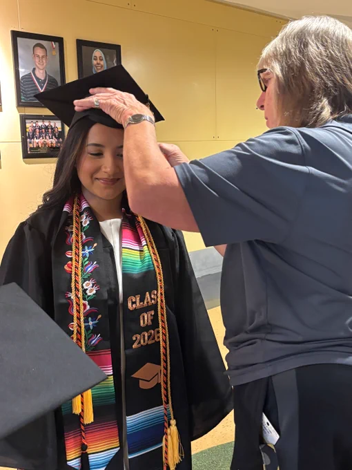 Graduate wearing a colorful stole and Class of 2026 sash having their cap adjusted by a staff member.