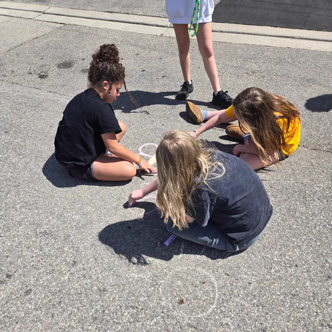 Three children sitting on asphalt drawing with chalk while a fourth child stands nearby on a sunny day.