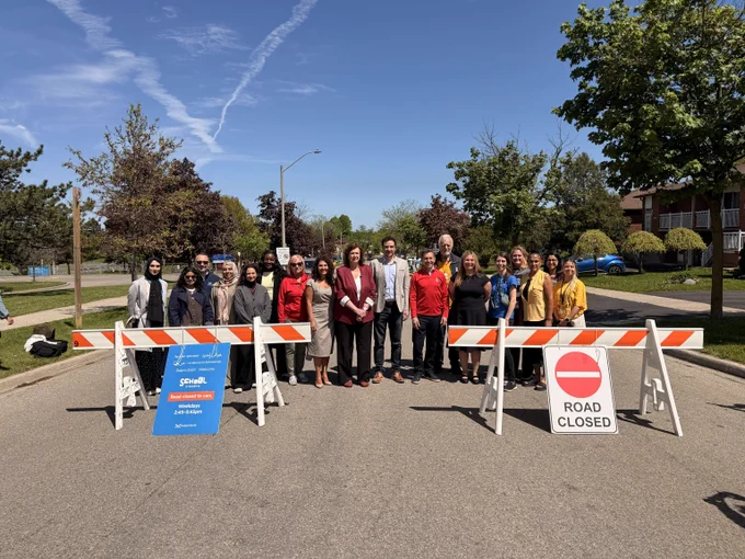 Group of people standing behind road closure barriers on a sunny suburban street with clear blue sky.