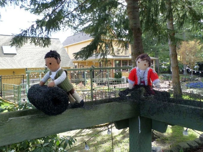 Crochet dolls of Cicero rolling the deer net along the twin arbor planks, while Edmund Hewlett gathers the excess on the inside of a 90 degree corner. Yard, garage & house in background. 