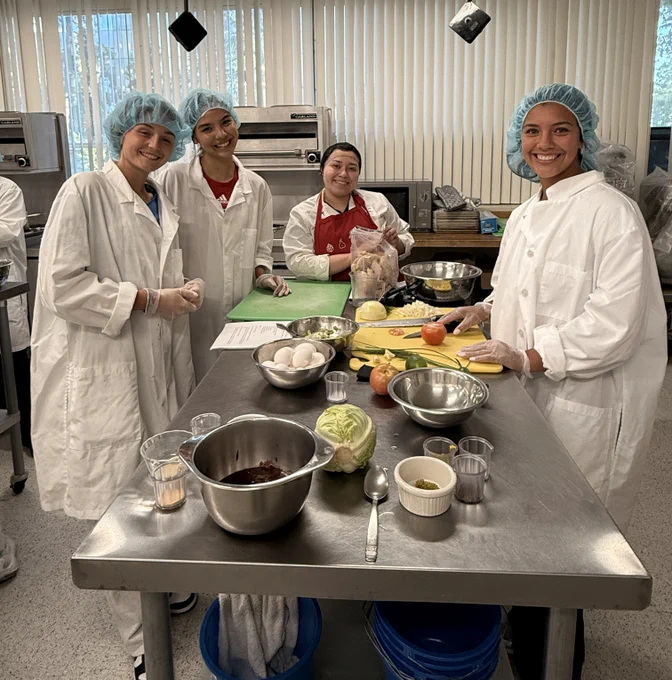 Four people are wearing lab coats and hairnets, standing around a stainless steel table in a kitchen setting. Various ingredients, including eggs, apples, lettuce, and spices, are on the table. They are smiling at the camera.
