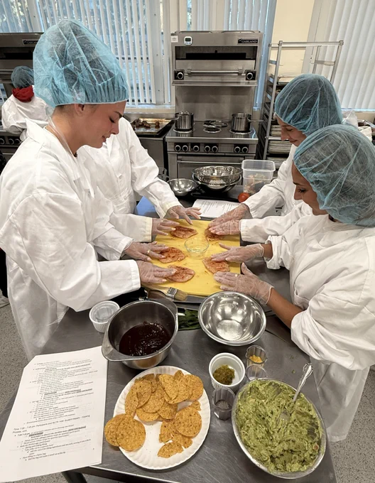 A group wearing hairnets and white coats is gathered around a table, shaping dough on a yellow surface in a kitchen. There are bowls with various ingredients, tortilla chips, and a dish of guacamole on the table. A recipe or instructions sheet is visible in the foreground.